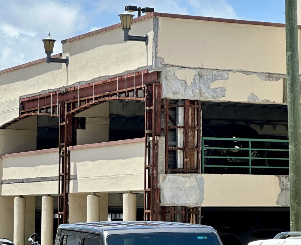 Large portions of stucco are missing from the exterior of Parking Garage No. 1 in Coral Gables, revealing rusted metal framework and signs of water damage. The visibly aged structure shows patchwork repairs and deterioration that raise both safety and aesthetic concerns.