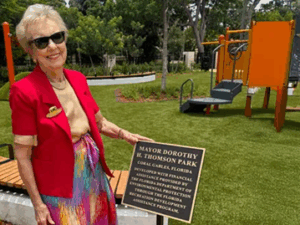 Dorothy Thomson, wearing sunglasses and a red jacket over a colorful skirt, smiles while holding the dedication plaque at the newly named Mayor Dorothy H. Thomson Park in Coral Gables.