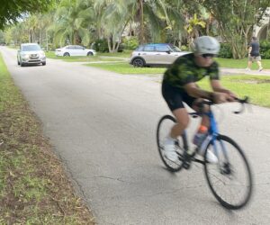 A cyclist in motion rides on a narrow residential street near Granada Golf Course, with a car approaching from behind and no sidewalk or bike lane visible.