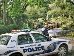 Coral Gables police officers and city workers stand near fallen trees and debris during post-Hurricane Irma cleanup in 2017, with a police car and heavy machinery visible in the scene.