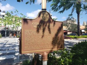 A bronze historical marker on Coral Gables’ Miracle Mile describes the street’s origins after World War II, with shops and palm trees in the background. The Gazette editorial argues that the city’s focus should be on the future, not the past.
