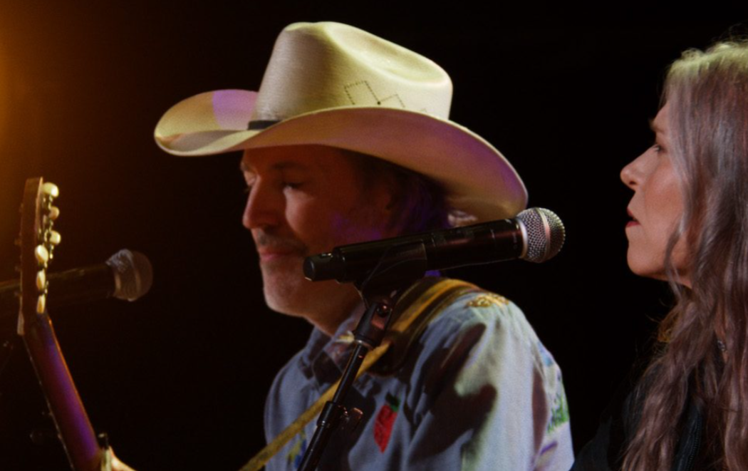 A musician in a light-colored cowboy hat plays acoustic guitar at a microphone while a female vocalist sings beside him during a live concert performance.
