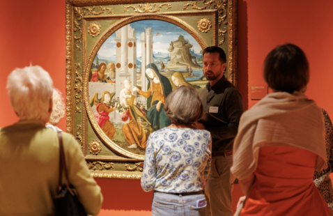 A museum docent speaks to a small group of visitors in front of a framed Renaissance painting at the Lowe Art Museum, explaining artistic details during an expert-led tour.