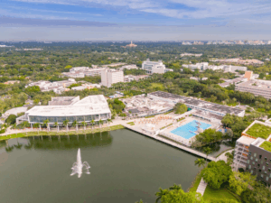 Aerial photograph of the University of Miami campus showing academic buildings, green spaces, a lake with a fountain, and athletic facilities surrounded by tree-lined neighborhoods.