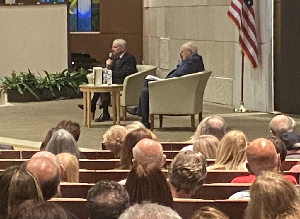 Jon Meacham speaking into a microphone while seated onstage beside moderator David Lawrence Jr. at Temple Beth Am in Pinecrest, addressing a packed audience during a Books & Books event.