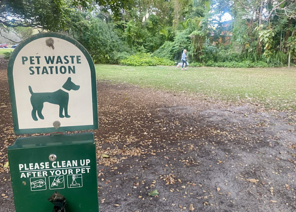 Pet waste station sign in a grassy park area with a person walking a dog in the background.
