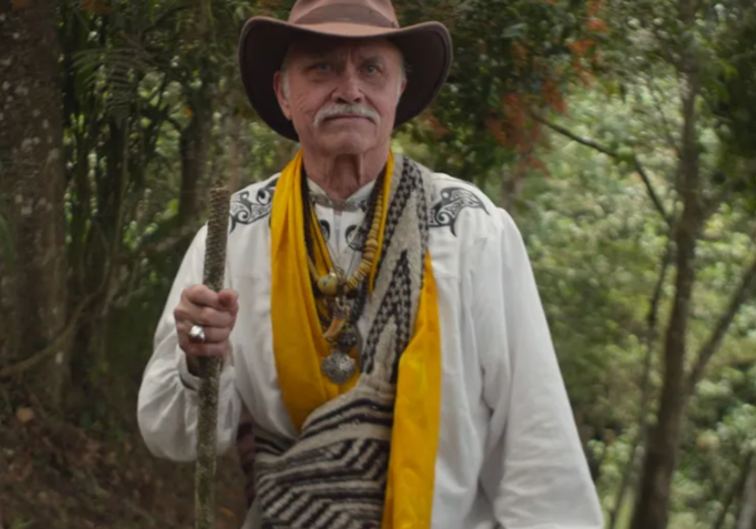 Patrick McCollum stands outdoors in a wooded setting, wearing a wide-brimmed hat, layered necklaces, and traditional-style garments, holding a walking stick, in a scene from the documentary The Man Who Saves the World?.