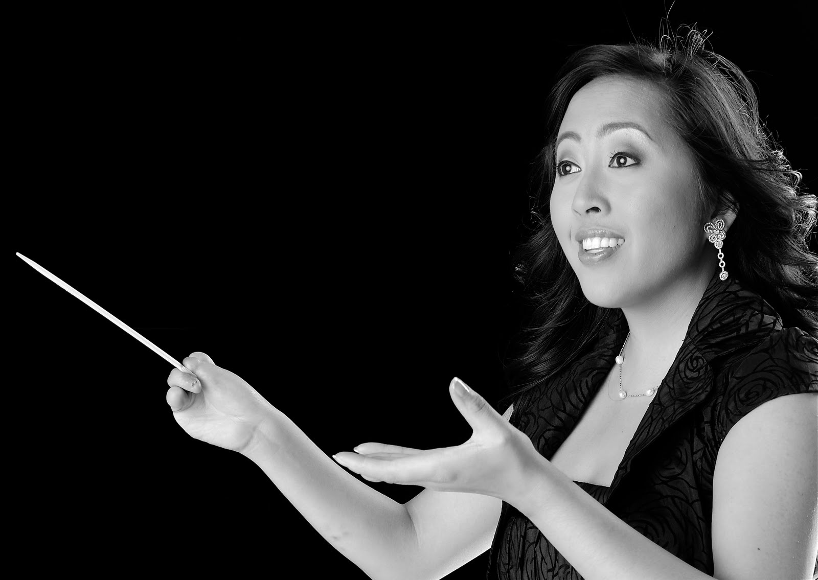 Black-and-white portrait of conductor Arianne Abela shown from the shoulders up against a solid black background. She holds a conductor's baton in her right hand and extends her left hand open, mid-gesture, with an expression of focused intensity. She wears a patterned sleeveless top, drop earrings, and a delicate pendant necklace.