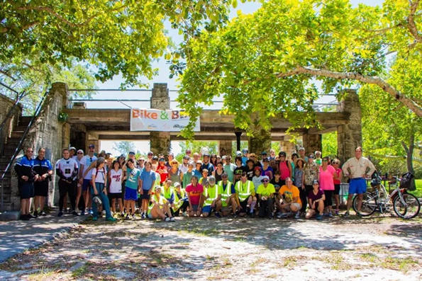 A large group of cyclists and walkers posed beneath a coral stone archway at Matheson Hammock Park, with bicycles visible at the edges of the crowd and trees overhead. A Bike & BBQ banner is visible in the background.