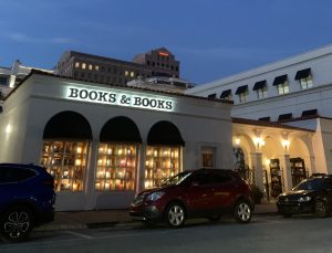 The exterior of Books & Books in Coral Gables photographed at dusk. The two-story white Mediterranean-style building is warmly lit from within, with arched colonnades and large windows displaying books. A illuminated sign reading "Books & Books" spans the roofline. Several cars are parked in front, and a multi-story building is visible in the background against a deep blue evening sky.