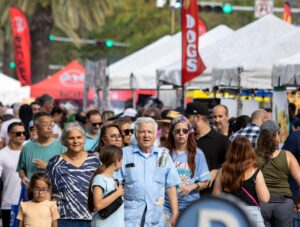 A dense crowd of multigenerational festival-goers walks along a closed street lined with vendor tents and promotional banners during the 2024 Carnaval on the Mile street festival in Coral Gables, Florida.