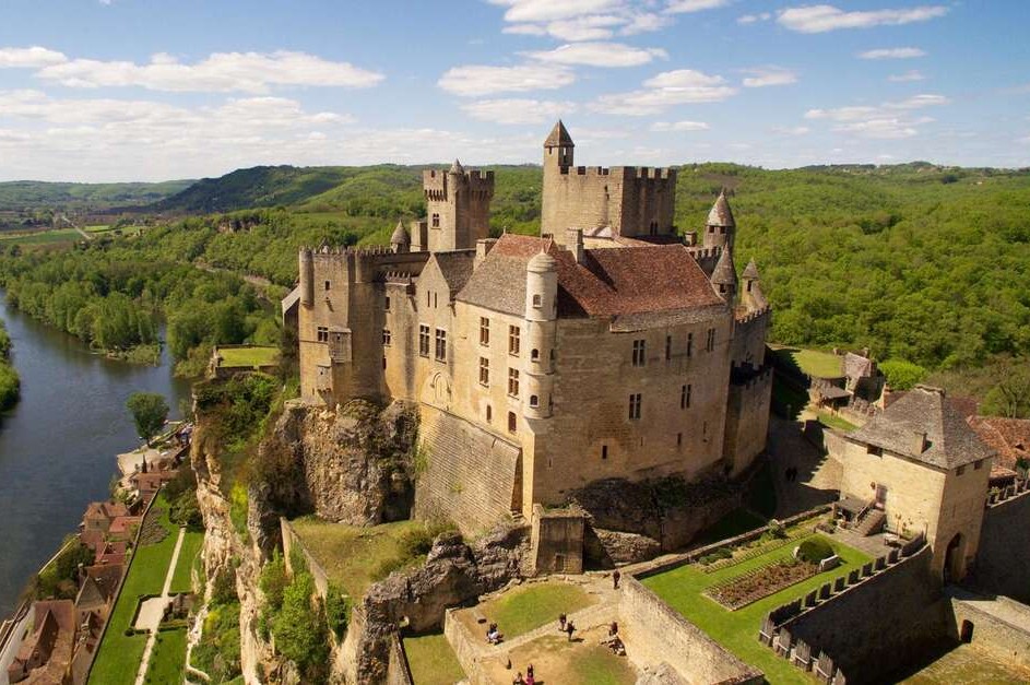 Aerial view of Château de Beynac, a large medieval stone fortress with multiple towers, crenellated battlements, and a red-tiled roof, perched on a steep limestone cliff above the Dordogne River. The surrounding landscape is dense green forest under a partly cloudy blue sky, with the river curving through the valley below and the rooftops of a small village visible at the base of the cliff.