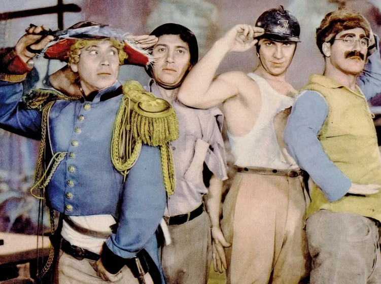 Four men in mismatched military costumes stand in a row, each saluting with one hand. From left: a man in a blue military jacket with gold buttons, a man in a tan shirt with gold-braided epaulettes, a man in a white undershirt wearing a small helmet, and a man in a yellow vest wearing round glasses and a false mustache. The background suggests a theatrical or battlefield setting with soft, colorful tones.
