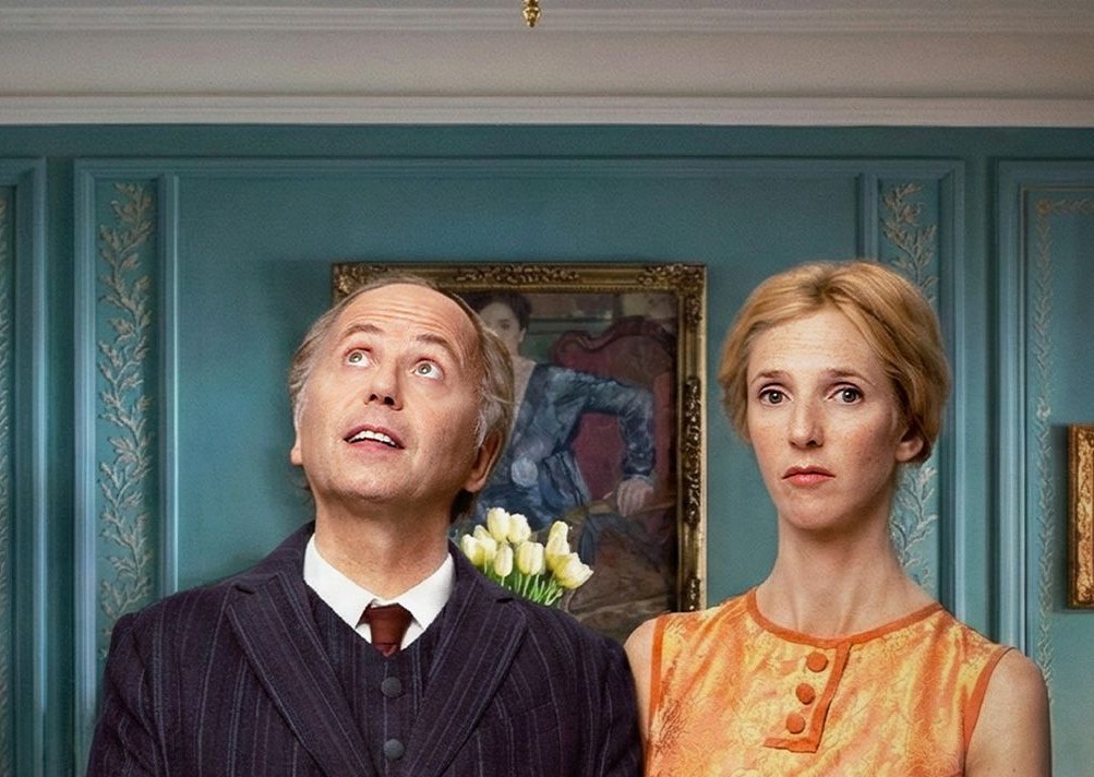 Two characters stand in an elegant Paris apartment, with the man looking upward and the woman facing forward, reflecting the film’s themes of class and social division.