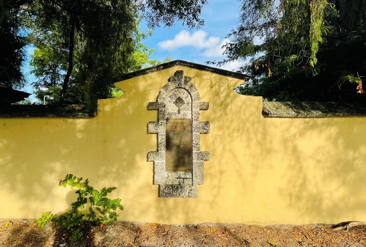 Memorial plaque and stone cross set into a yellow stucco wall at the Garden of Our Lord meditation garden in Coral Gables, surrounded by trees and greenery.