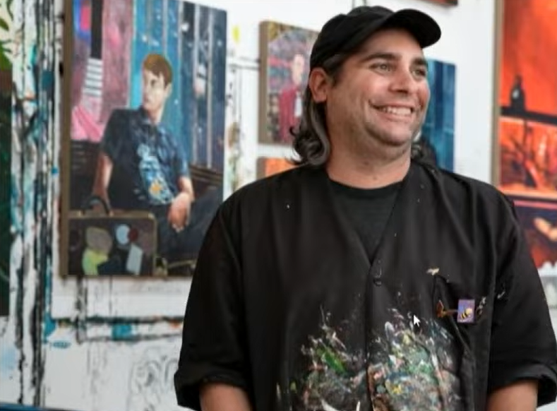Contemporary artist Hernan Bas stands smiling in his studio, wearing a paint-splattered smock and cap, with several colorful figurative paintings displayed on the walls behind him.