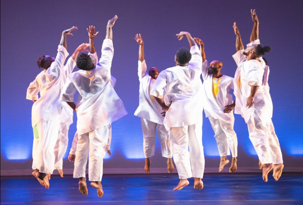 Group of contemporary dancers in white costumes leaping with arms raised on a stage lit in blue and purple tones.