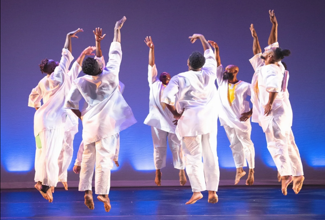 Group of contemporary dancers in white costumes leaping with arms raised on a stage lit in blue and purple tones.
