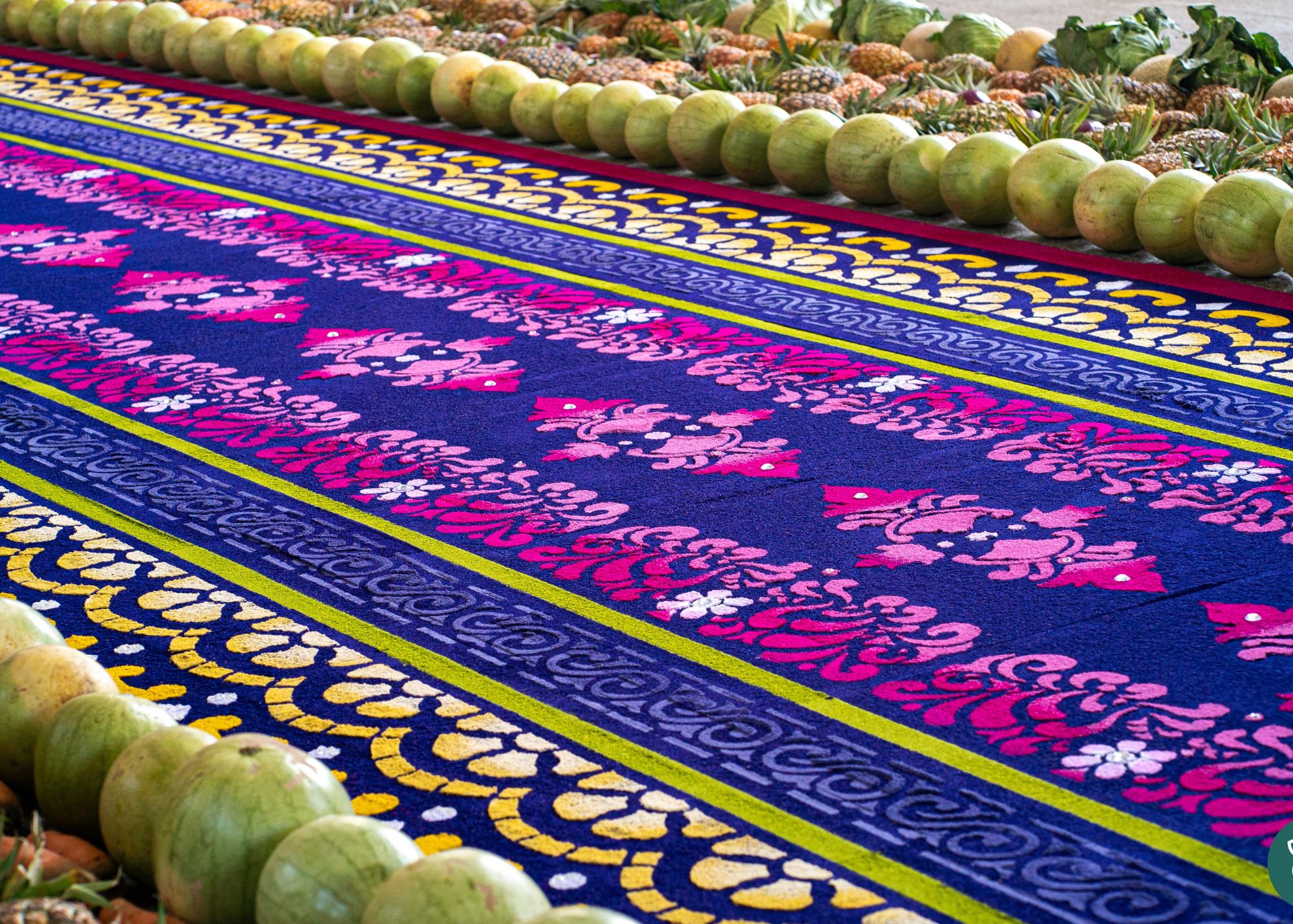 A brightly colored sawdust carpet featuring intricate geometric and floral patterns is arranged on the ground, bordered by rows of green fruit and other natural materials.