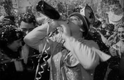 A black-and-white film still from "I Vitelloni" showing a man in a carnival costume drinking directly from a large bottle amid a crowd of revelers. Confetti, streamers, and festive decorations fill the frame, and other costumed figures are visible in the background.