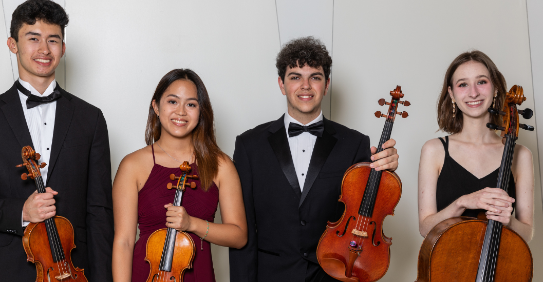 Four young musicians in formal attire stand in a row against a white wall, each holding their instrument. From left: a young man in a tuxedo holding a violin; a young woman in a burgundy gown holding a violin; a young man in a black suit and bow tie holding a viola; and a young woman in a black dress holding a cello. All four are smiling.
