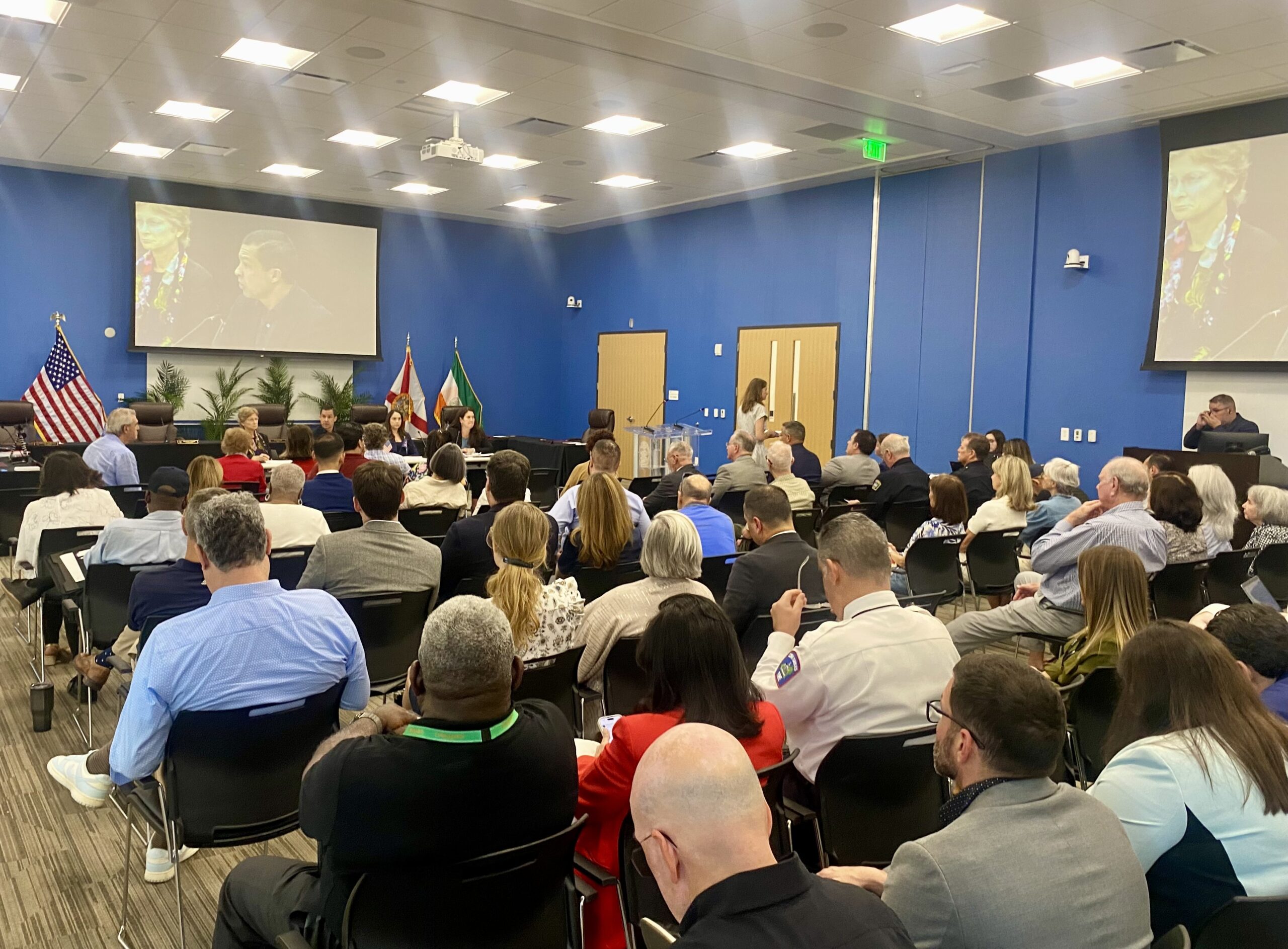 A packed community meeting room at the Coral Gables Public Safety Building, with rows of residents seated facing a panel of city officials at a front table, American and Florida flags flanking the dais, and two large projection screens displaying a video presentation on the blue walls.