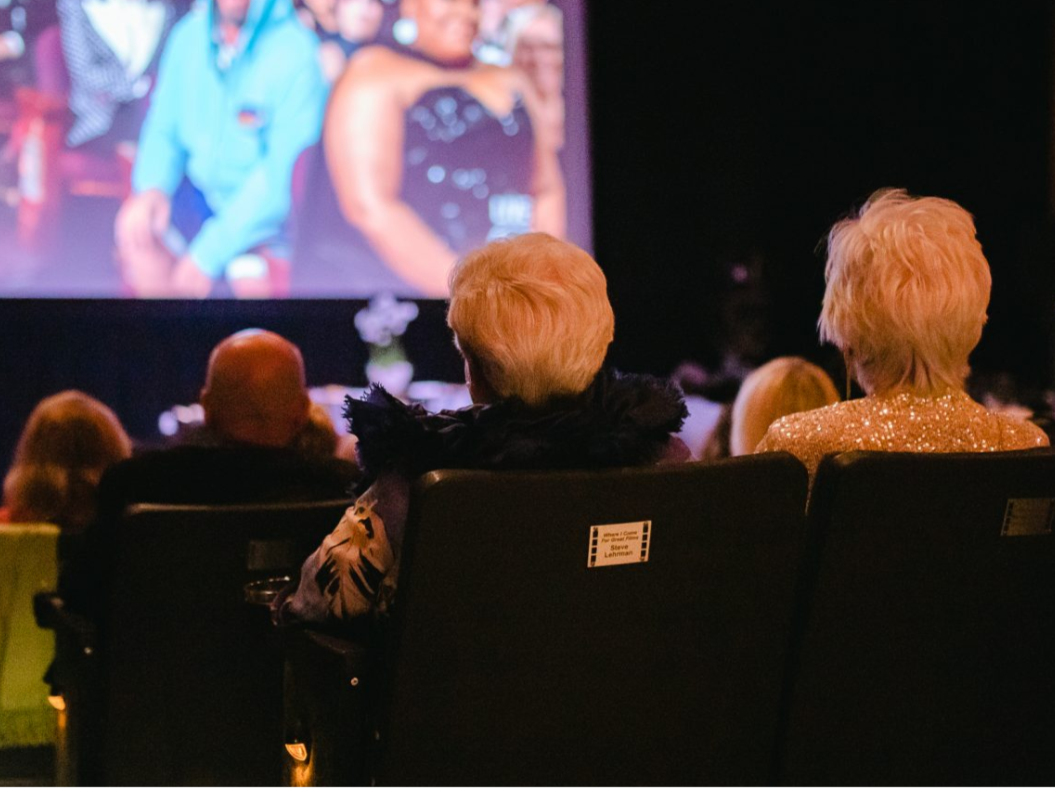 Audience members seated in a darkened cinema theater face a large illuminated screen showing awards broadcast footage, with several guests dressed in formal and sequined attire visible from behind.