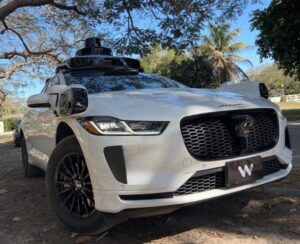 White Waymo Jaguar I-Pace autonomous vehicle equipped with roof-mounted sensors parked along a tree-lined residential street in Coral Gables.