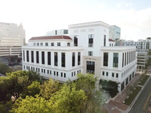 Aerial view of the Coral Gables Public Safety Building, a large white multi-story structure with a Mediterranean-influenced facade, red tile roof accents, black-framed windows, and a central entrance marked with the city and department name in stone lettering. The building is surrounded by mature tropical trees and situated in an urban downtown context with commercial and residential buildings visible in the background.