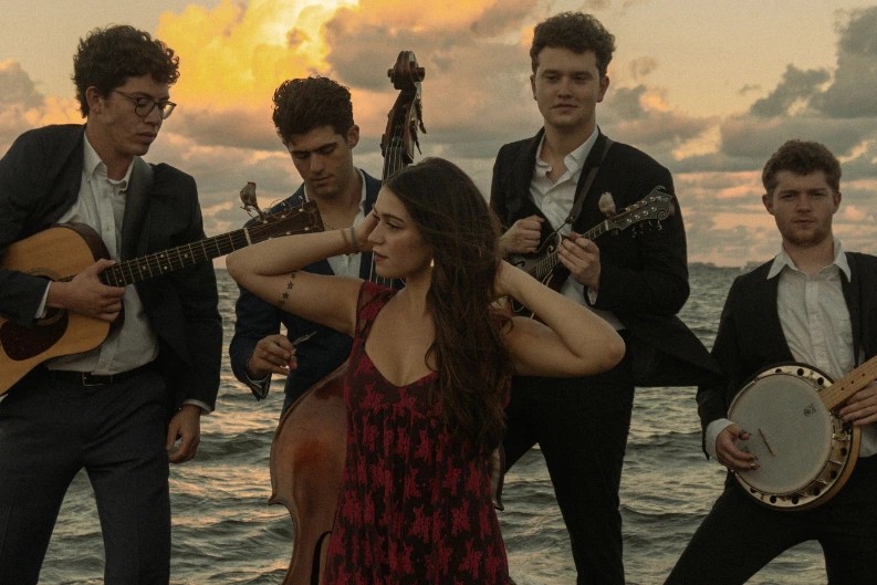 Five musicians perform with acoustic instruments, including banjo, mandolin, guitar, and upright bass, standing by the water at sunset with a female vocalist in the foreground.