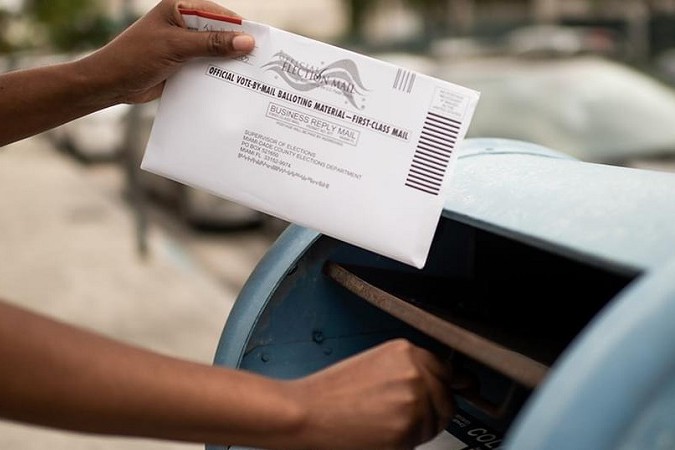 A person's hands hold a white official vote-by-mail balloting material envelope addressed to the Miami-Dade County Supervisor of Elections over the open slot of a blue outdoor USPS mailbox, preparing to drop it in. Sonnet 4.6
