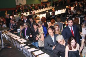 A crowded indoor event shows people in formal attire gathered around tables reviewing auction items and bidding sheets.