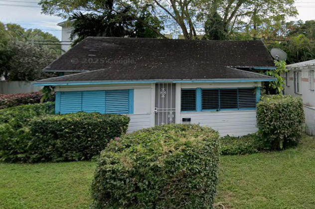 A small single-story house with a sloped roof, light-colored exterior, blue shutters, and a front yard with trimmed hedges and grass.