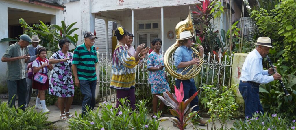 Street scene with musicians playing clarinet and tuba as residents walk and clap in a Cuban neighborhood.