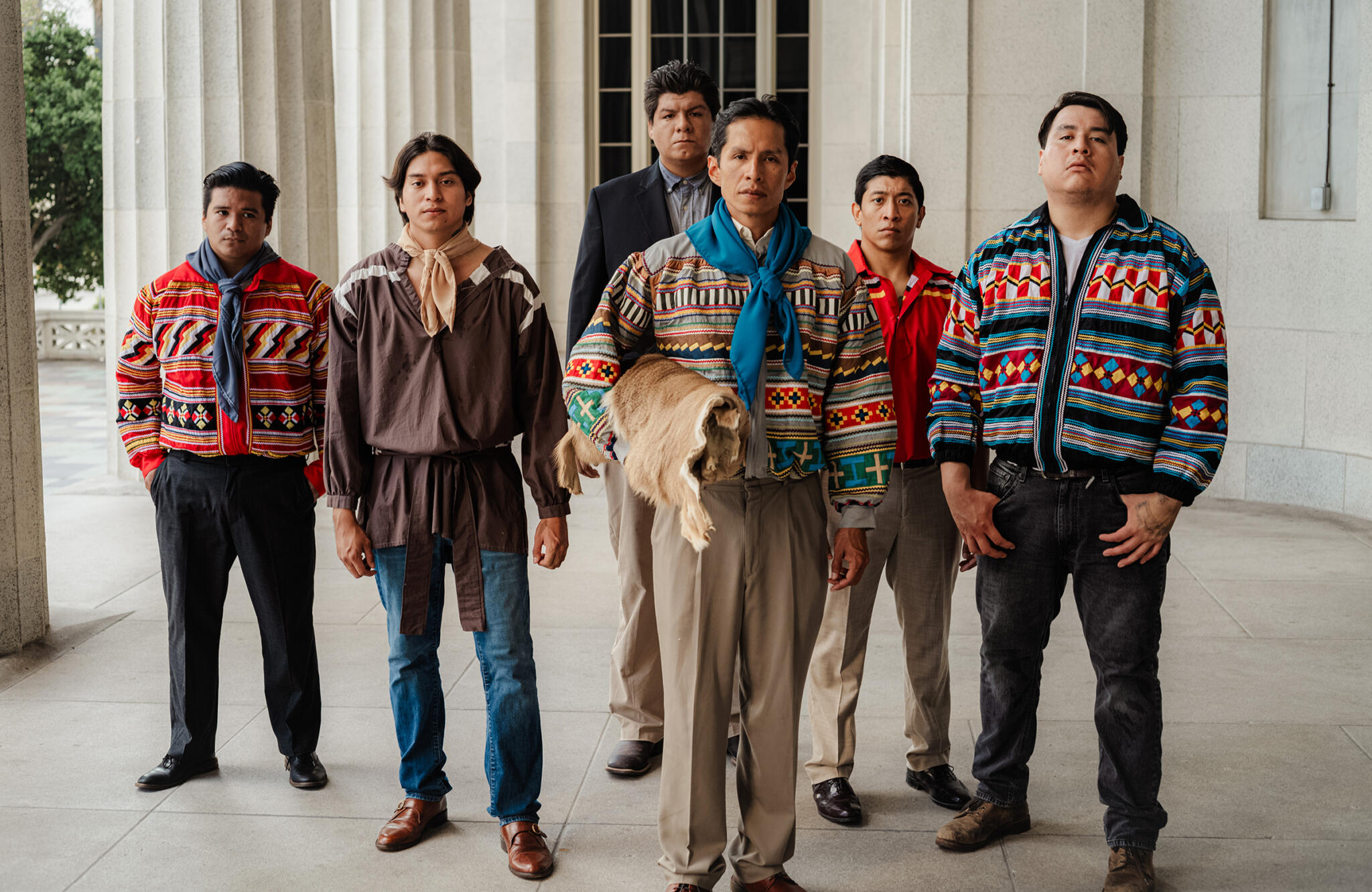 A production still from the film Becoming Buffalo. Six men stand in a formal row before a building with large white marble columns, suggesting a government or institutional setting. The man at the center foreground wears traditional Miccosukee patchwork clothing — a brightly colored jacket with geometric patterns in red, green, white, and teal — with a turquoise scarf around his neck and holds a folded animal hide. The other men wear variations of traditional Miccosukee patchwork jackets in bold multicolor geometric designs, with the exception of one man in the background who wears a dark suit. All six look directly at the camera with composed, serious expressions. The contrast between the vibrant traditional garments and the formal stone architecture behind them is visually striking.