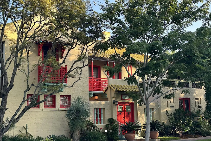A two-story stucco home in the Chinese Village of Coral Gables, photographed in late afternoon light. The exterior is painted pale yellow with vivid red architectural details throughout — a latticed upper balcony with classical Chinese fretwork patterns, red-trimmed windows with geometric grille inserts, a bold red front door with a decorative brass lantern overhead, and red accent trim along the roofline. A green ceramic tile roof with slightly upturned eaves is visible at the top of the frame. Mature trees frame both sides of the facade, and tropical plantings including a yucca and potted succulents border the entrance. The house number 5133 is visible beside the front door.