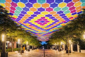Colorful crocheted canopy suspended above a pedestrian street lined with trees and storefronts at night, forming a patterned overhead installation at Giralda Plaza.