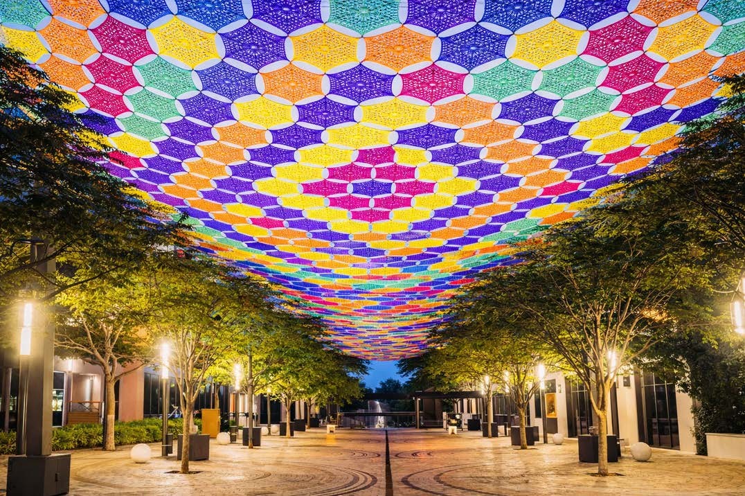 Colorful crocheted canopy suspended above a pedestrian street lined with trees and storefronts at night, forming a patterned overhead installation at Giralda Plaza.