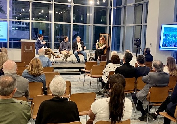 A panel discussion in a modern, glass-walled room at the University of Miami. Four panelists are seated on a low stage facing an audience of approximately 50 people seated in wooden chairs. From left, a man in a dark suit speaks into a microphone at a podium; two men in casual attire sit in chairs on stage; and a woman in a black-and-white patterned top holds a microphone. A projection screen is visible to the right. The audience is seen from behind, engaged and attentive. The room is lit warmly against a darkened exterior visible through the large windows.
