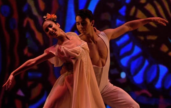 Two ballet dancers perform on a dramatically lit stage against a vivid abstract backdrop in deep blues, purples, and reds. The female dancer is in the foreground, wearing a flowing sheer white dress with an orange flower in her hair, leaning back with her left arm extended gracefully. The male dancer is positioned behind and to her right, his right arm extended fully outward, wearing a white sleeveless costume. Both performers are mid-movement, their expressions focused and expressive.