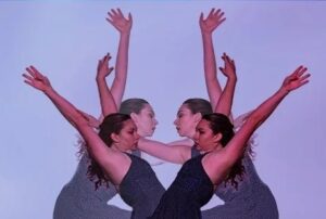 Three dancers arranged in a mirrored composition extend their arms upward and outward against a soft purple background, creating overlapping silhouettes and a symmetrical visual effect.