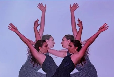 Three dancers arranged in a mirrored composition extend their arms upward and outward against a soft purple background, creating overlapping silhouettes and a symmetrical visual effect.