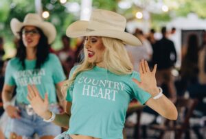 A line dancing instructor in the foreground wears a cream cowboy hat and a teal Country Heart t-shirt, arms raised and mouth open mid-call, leading a dance session at an outdoor evening event. String lights glow overhead against a backdrop of lush green trees. A second instructor in an identical outfit and cowboy hat is visible slightly behind and to the left. A crowd of attendees is blurred in the background. The setting suggests a warm outdoor evening at Fairchild Tropical Botanic Garden.