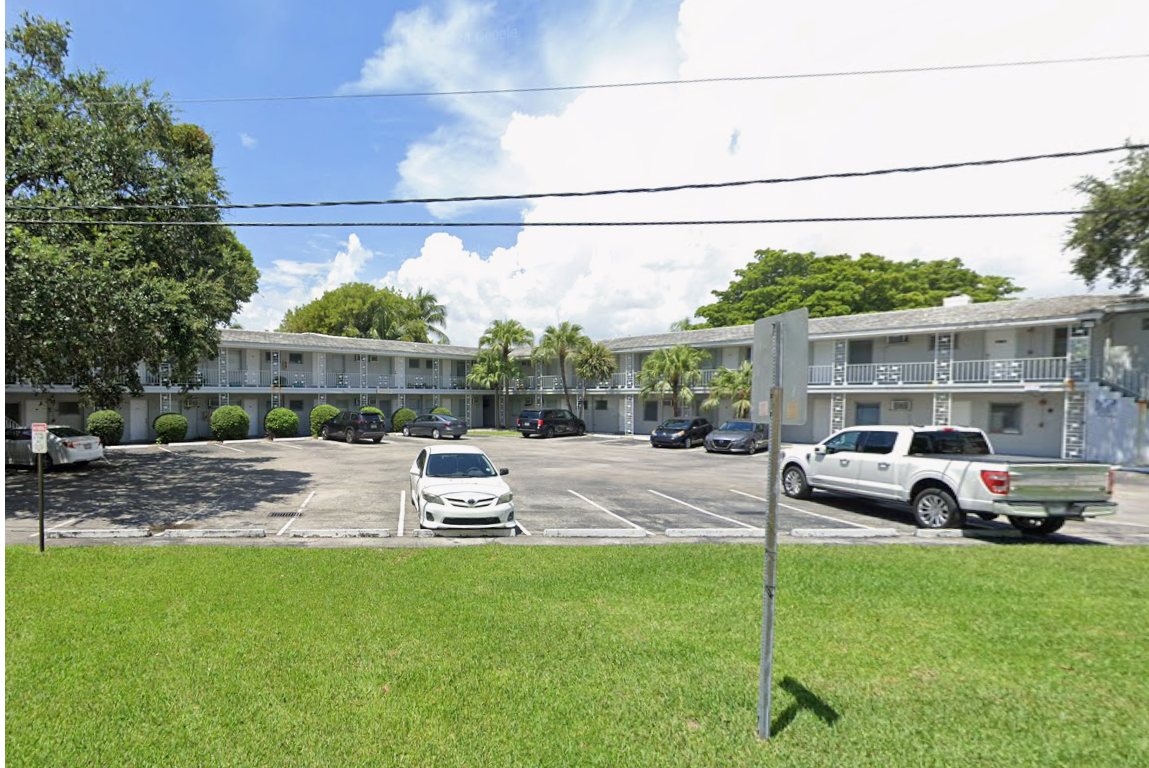 Street-level view of a two-story apartment complex at 6100 Caballero Boulevard in Coral Gables. The building has a mid-century utilitarian design with exterior walkways, metal railings, and window air conditioning units. Several cars are parked in a surface lot in front of the building. Mature trees and a manicured lawn border the property. Power lines run across the upper portion of the image.