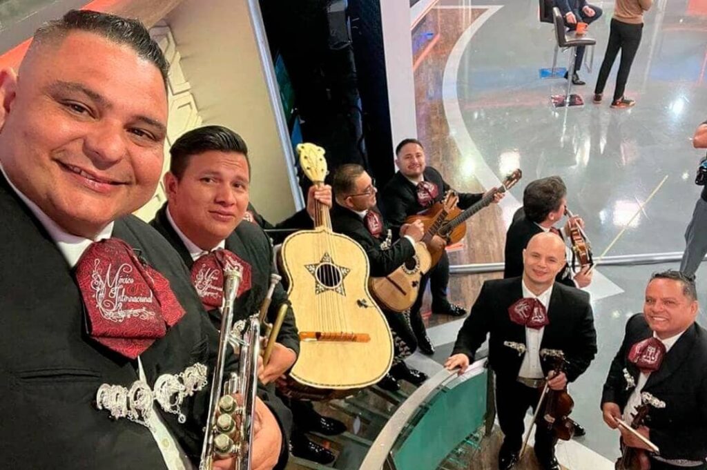 Six members of Mariachi México Internacional pose for a selfie in matching black charro suits with burgundy embroidered pocket squares. In the foreground, a trumpet and a vihuela are visible; behind them, musicians hold a guitarrón, a guitar, and two violins.