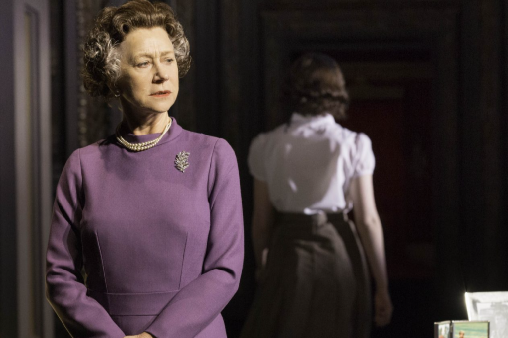 A scene from the stage production of The Audience. In the foreground, a woman portraying Queen Elizabeth II stands facing slightly left, wearing a purple long-sleeved dress with a pearl necklace and silver brooch. Her gray hair is styled in a period-appropriate set. Her expression is composed and watchful. In the background, a second woman in a white blouse and dark skirt stands with her back to the camera in a dimly lit, wood-paneled interior setting.