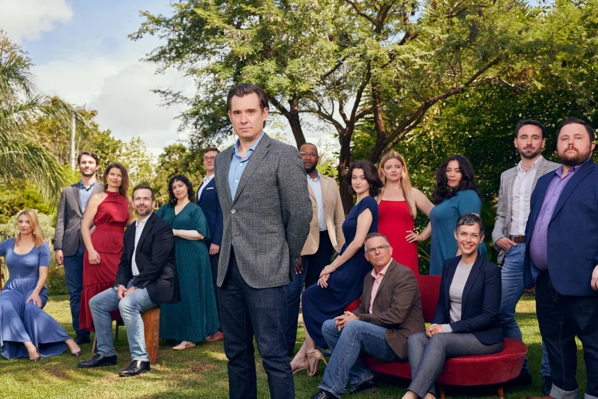 A group portrait of Patrick Dupré Quigley and members of the Seraphic Fire ensemble, photographed outdoors against a backdrop of lush tropical trees and a partly cloudy sky. Quigley stands in the foreground, centered, wearing a gray textured blazer and light blue shirt, looking directly at the camera with a composed expression. Behind and around him, approximately 14 ensemble members are arranged in a loose grouping — some standing, some seated on a red velvet sofa — dressed in a mix of formal and smart casual attire in colors including navy, red, teal, blue, and gray. The setting appears to be a garden or landscaped outdoor space in South Florida.