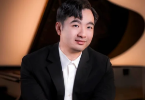 Portrait of pianist William Yang seated in front of a grand piano, wearing a black suit and white shirt.