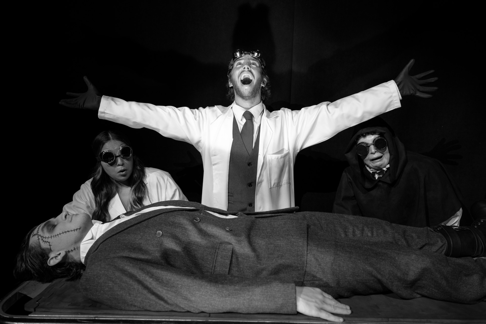 A black-and-white stage scene shows a lab setting with a man in a white coat raising his arms over a figure lying on a table, while two other characters in goggles stand nearby.