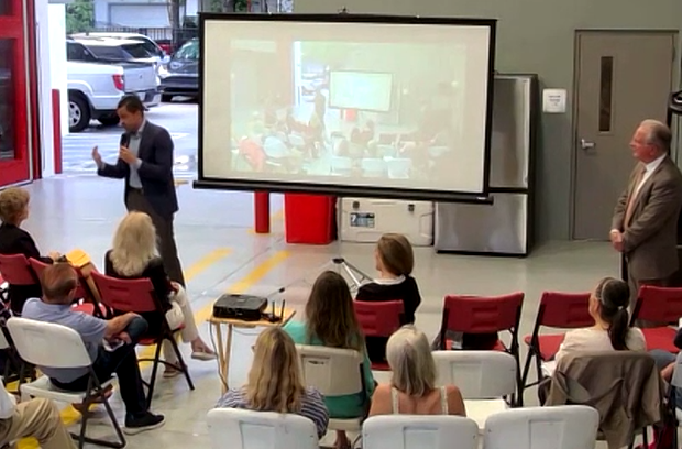 Mayor Vince Lago stands at left holding a microphone and gesturing toward the audience inside the bay of Firehouse 4 on Sunset Drive. A large projection screen behind him displays a live Zoom feed of meeting participants joining remotely. City Manager Peter Iglesias stands at right in a gray suit, hands clasped, observing. Residents seated in red chairs face the presenters with their backs to the camera. The garage bay door is open behind the officials, with a vehicle and evening light visible outside. A projector sits on a small table in the center of the room.